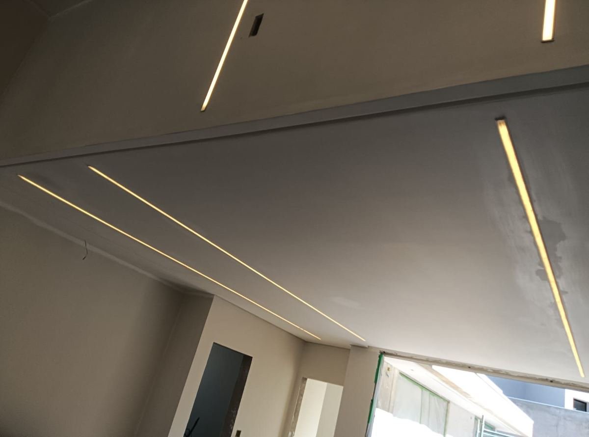 Forro de gesso com recortes lineares de luz embutida acesos, acompanhando as quebras do teto de um ambiente em acabamento, instalados pela J&C Elétrica.