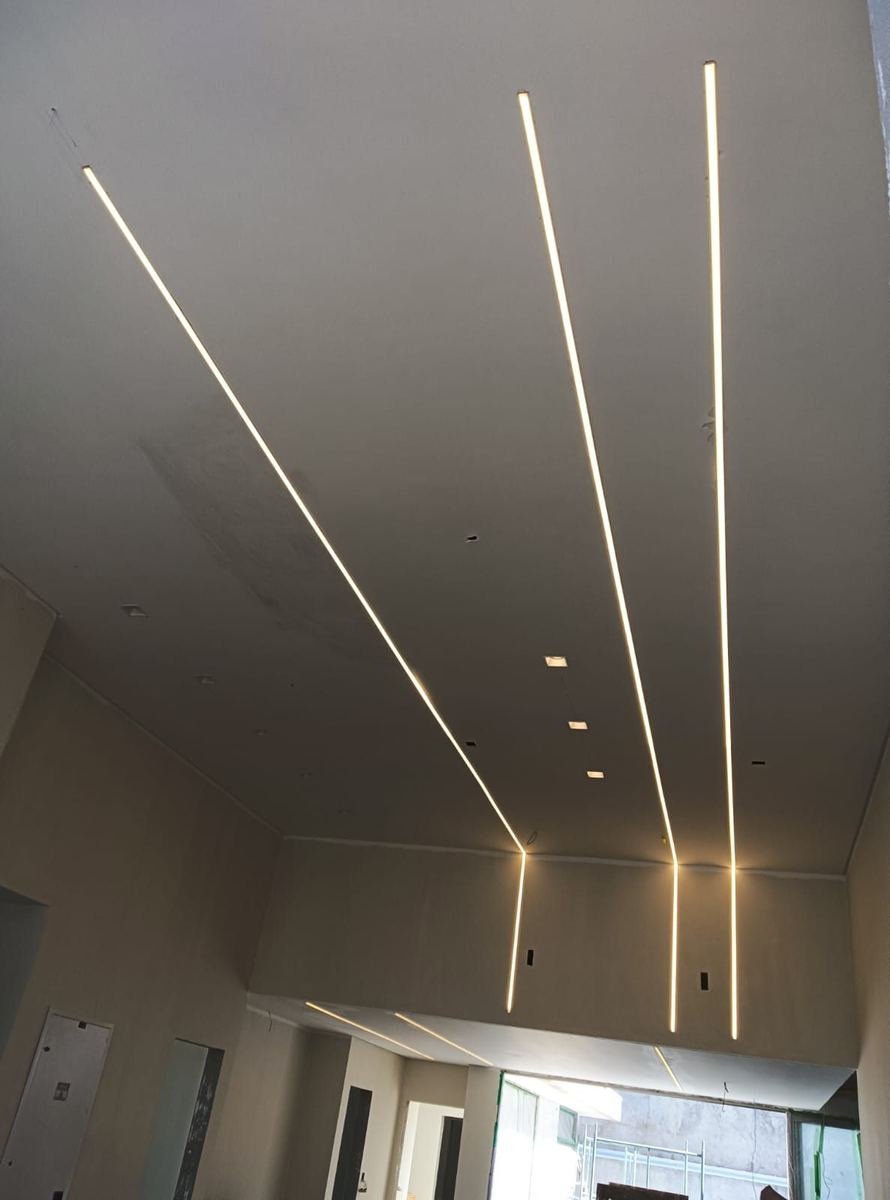 Teto alto com quatro luminárias lineares embutidas acesas, formando linhas de luz contínuas que atravessam o ambiente, instaladas pela J&C Elétrica.