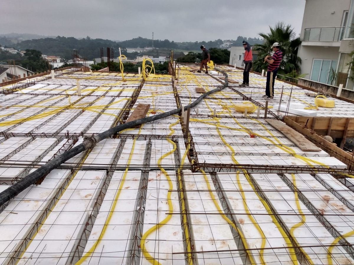 Vista ampla de uma laje pré-moldada armada, com eletrodutos amarelos percorrendo toda a extensão e a equipe trabalhando ao fundo, obra da J&C Elétrica.