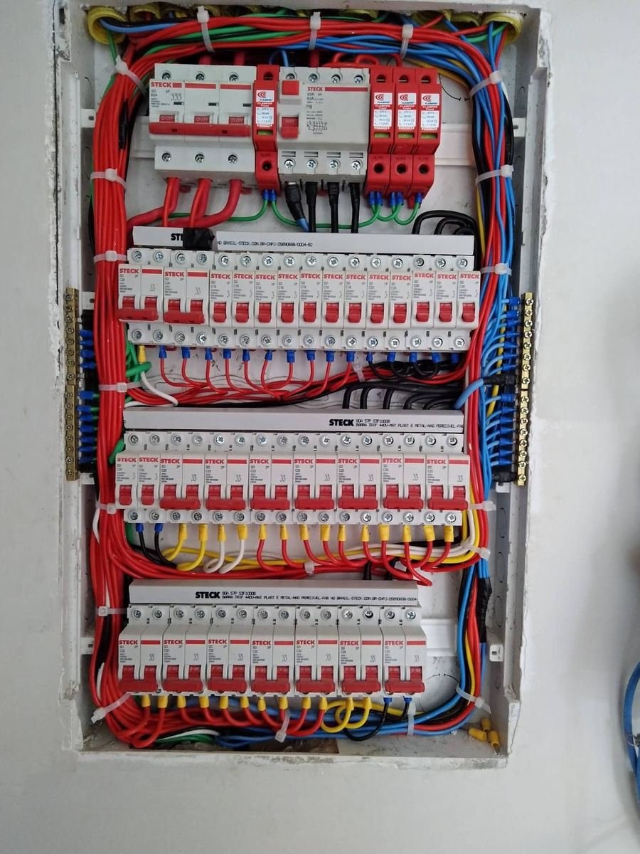 Quadro de distribuição com três fileiras de disjuntores sobre barramentos, cabos vermelhos e azuis organizados e amarrados, protetores contra surto na entrada, montado pela J&C Elétrica.